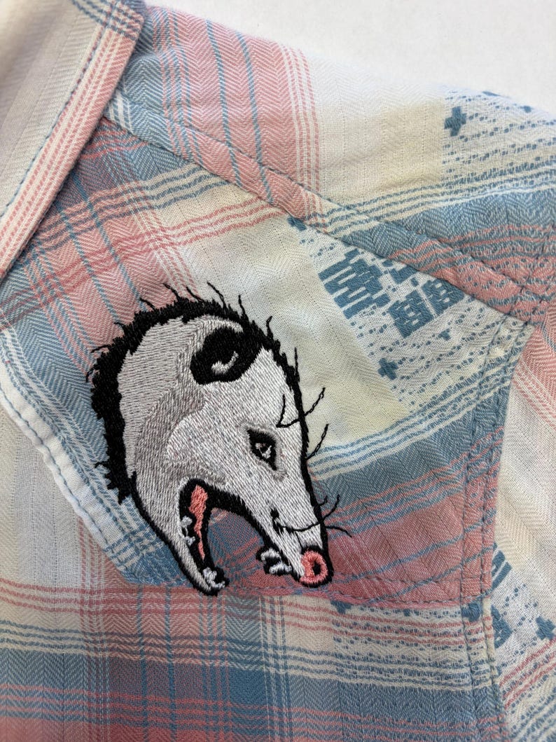 May include: Close-up of a plaid shirt in pastel pink, blue, and white. The shirt features an embroidered opossum patch on the pocket. The opossum is gray and white with a pink mouth and black accents.