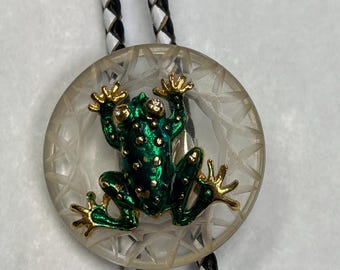May include: A bolo tie with a green frog and gold detailing on a clear, faceted disc. The frog has a speckled texture. The tie's cord is made of black and white braided leather.
