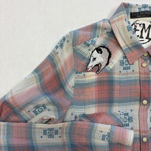 May include: A button-down plaid shirt in shades of pink, blue, and white. An embroidered opossum patch is on the chest. The shirt has a collar and long sleeves with buttoned cuffs. The label reads "Ellie Monsters Nashville".