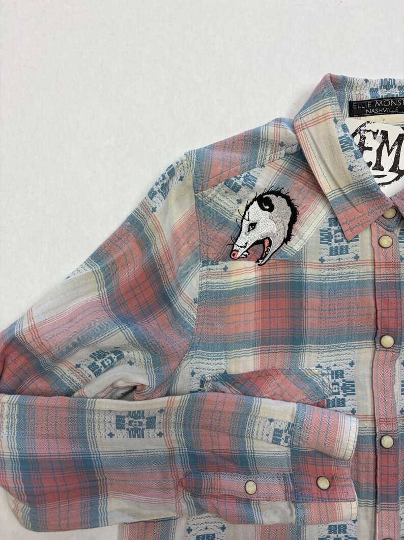 May include: A button-down plaid shirt in shades of pink, blue, and white. An embroidered opossum patch is on the chest. The shirt has a collar and long sleeves with buttoned cuffs. The label reads "Ellie Monsters Nashville".