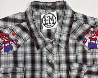 May include: A long-sleeve button-down shirt in a grey, black, and white plaid pattern. The shirt features embroidered clown figures on both shoulders and a collar tag reading Ellie Monster Nashville with a white tag displaying the letters EM.