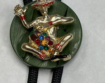 May include: A gold-coloured bolo tie featuring a seated figure with a red hat and accents. The figure is adorned with colourful gemstones and sits on a green circular base. The tie has a black braided cord.