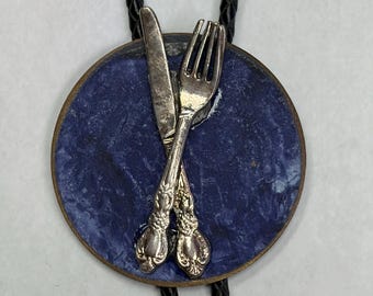 May include: A bolo tie featuring a circular pendant with a deep blue, marbled surface. Attached to the centre of the disc are two miniature silver-toned utensils, a knife and a fork, crossed in an X shape. The pendant hangs from a black braided cord.