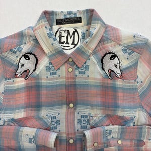 May include: A button-down plaid shirt in pink, blue, and white with two opossum patches on the chest. The shirt has a collar and cuffs with pearl snap closures. The label reads "Ellie Monster Nashville".