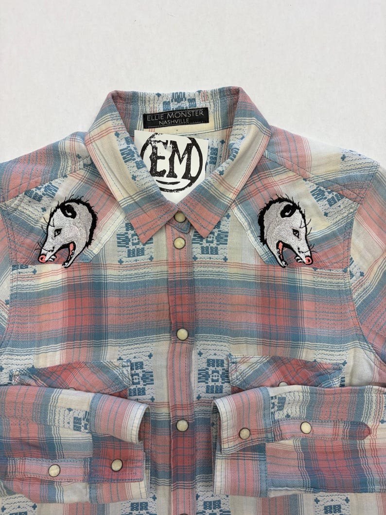 May include: A button-down plaid shirt in pink, blue, and white with two opossum patches on the chest. The shirt has a collar and cuffs with pearl snap closures. The label reads "Ellie Monster Nashville".