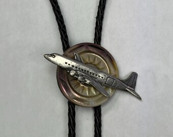 May include: A bolo tie featuring a silver-toned metal aeroplane charm mounted on a circular, iridescent, brown-toned button. The charm is attached to a black braided leather cord that extends upwards and downwards from the central button piece.