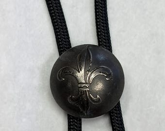 May include: A silver-toned bolo tie with a round, oxidized metal slide, displaying a fleur-de-lis design. The slide is attached to a black braided cord. A classic accessory.