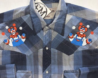 May include: Blue plaid shirt with embroidered clown motifs. The shirt has a collared neckline, two chest pockets, and snap closures. The tag reads "Ellie Monster Nashville". The shirt is a classic Western style.