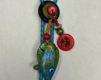 May include: A bolo tie featuring a braided light blue cord and a central focal point composed of a dark purple circular base, a green disc, and a small pink bead. A hand-painted blue and green fish charm hangs alongside a red disc and beads.