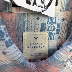 May include: Close-up of a Vintage Boyfriend shirt from American Eagle Outfitters. The shirt features a pattern of blue, pink, and white geometric designs. The label reads "Vintage Boyfriend" and "American Eagle Outfitters."