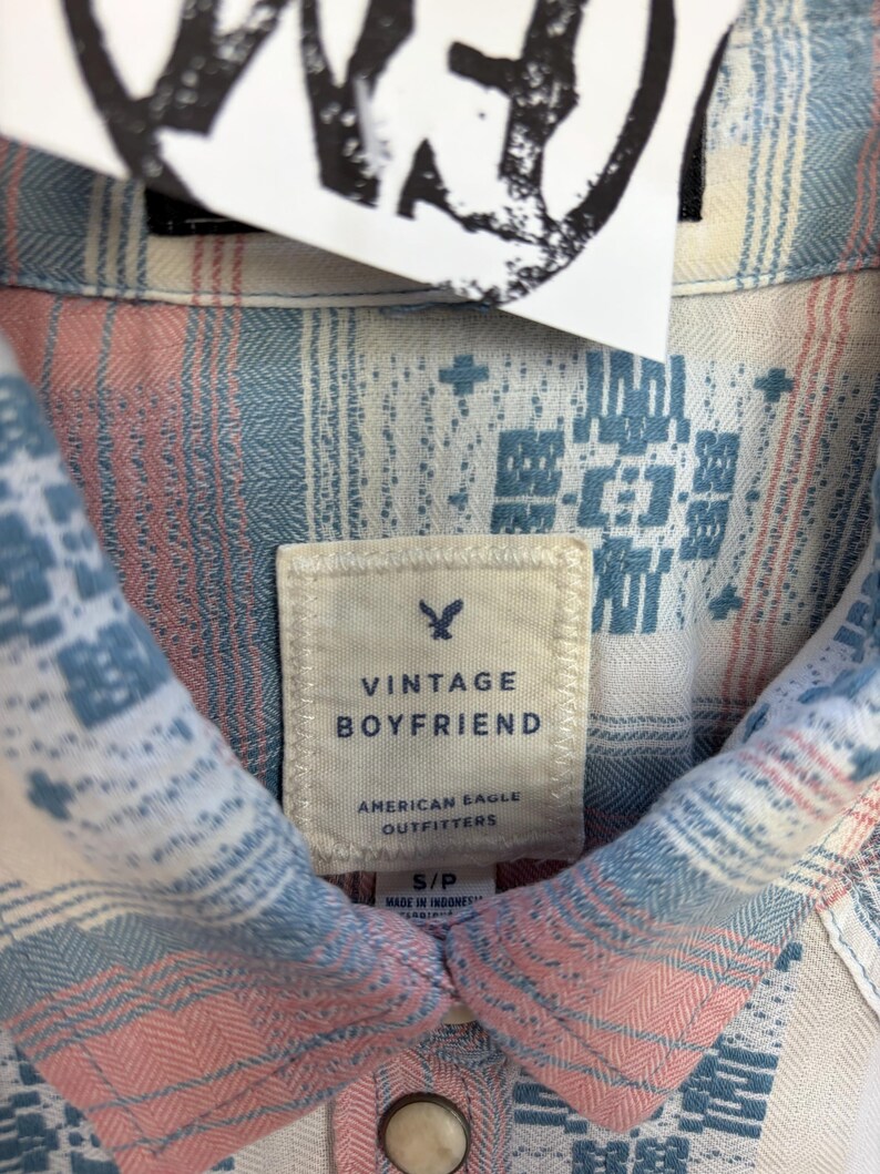 May include: Close-up of a Vintage Boyfriend shirt from American Eagle Outfitters. The shirt features a pattern of blue, pink, and white geometric designs. The label reads "Vintage Boyfriend" and "American Eagle Outfitters."