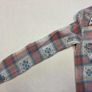 May include: A long-sleeved plaid shirt in shades of pink, blue, and white. The shirt features a small pocket and a button closure. An embroidered opossum patch is on the upper chest. The plaid pattern includes geometric designs.