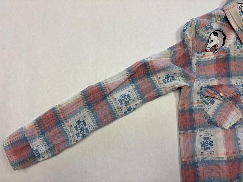 May include: A long-sleeved plaid shirt in shades of pink, blue, and white. The shirt features a small pocket and a button closure. An embroidered opossum patch is on the upper chest. The plaid pattern includes geometric designs.