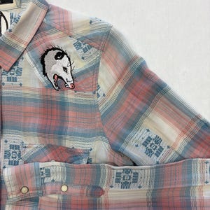 May include: A button-down shirt with a plaid pattern in shades of pink, blue, and cream. The shirt features an embroidered opossum patch on the chest. The sleeves are cuffed, and the shirt has two front pockets.