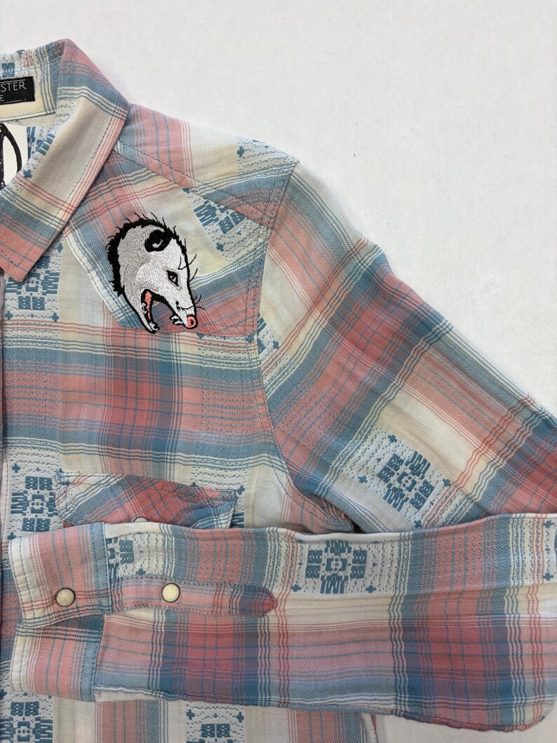 May include: A button-down shirt with a plaid pattern in shades of pink, blue, and cream. The shirt features an embroidered opossum patch on the chest. The sleeves are cuffed, and the shirt has two front pockets.