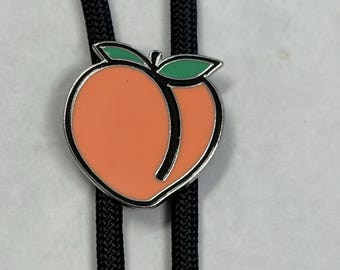 May include: A peach-shaped bolo tie pendant with an orange centre, black outline, and green leaves. The pendant is attached to a black braided cord, creating a unique accessory.