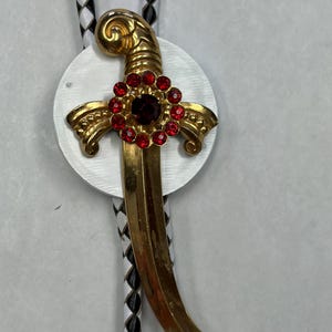 May include: A gold-colored bolo tie featuring a decorative dagger design with red gemstone accents. The dagger is set against a white circular backdrop, with a black and white braided cord.