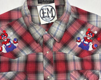 May include: A red, white, and black plaid button-down shirt featuring embroidered clown figures on the chest. The shirt has a collar, two chest pockets with snap closures, and a label reading Ellie Monster Nashville with a logo tag.