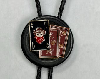 May include: A bolo tie featuring a black circular base with a braided black cord. The centerpiece is an enamel pin depicting three playing cards, including an ace of spades with a skull wearing a cowboy hat and red bandana, and two red cards.