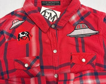 May include: Red plaid shirt with UFO and cow patches. The shirt features a button-down front, two chest pockets, and the label "ELLIE MONSTER NASHVILLE".
