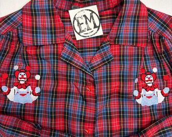 May include: A long-sleeve button-down shirt in a red, blue, and black plaid pattern. The shirt features two embroidered patches of a clown juggling balls on the chest. A white tag with the black letters EM in a circle is attached at the collar.