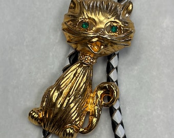 May include: A gold-coloured cat bolo tie with green gem eyes. The cat is sitting with its tail curled. The bolo tie cord is black and white braided leather.