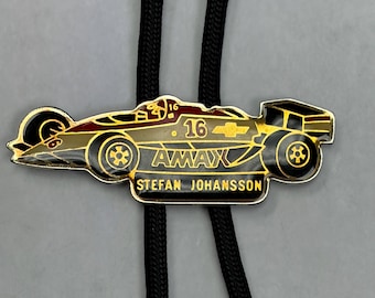 May include: A bolo tie featuring a gold-toned metal racing car pendant with black and red enamel details. The car is marked with the number 16, the text AMAX, and the name STEFAN JOHANSSON. It is attached to a black braided cord necklace.