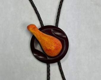 May include: A bolo tie featuring a dark brown circular slide with a textured orange and yellow chicken drumstick figure attached to the centre. The cord is made of black braided material that extends upwards and downwards from the slide.