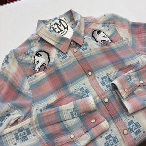 May include: A button-down plaid shirt in pink, white, and blue, featuring embroidered opossum designs on the chest. The shirt has a Western style with snap closures and decorative patterns on the cuffs and yoke. A tag with the letters "EM" is attached.