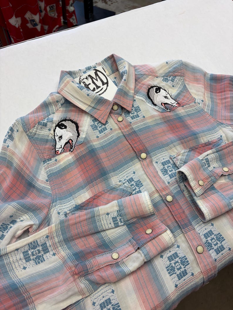 May include: A button-down plaid shirt in pink, white, and blue, featuring embroidered opossum designs on the chest. The shirt has a Western style with snap closures and decorative patterns on the cuffs and yoke. A tag with the letters "EM" is attached.