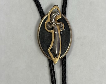 May include: A bolo tie featuring a black braided cord and an oval-shaped slide. The slide has a black centre inlay surrounded by a gold-toned metal border, with a silver-toned metal sword emblem centred on the front of the oval piece.