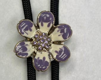 May include: A floral bolo tie with a flower-shaped pendant. The pendant has cream and purple enamel petals, a gold-tone border, and a cluster of small purple rhinestones. The tie has a black braided cord.