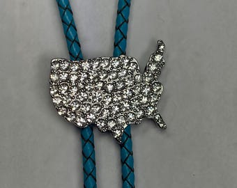 May include: A bolo tie featuring a silver-toned metal pendant shaped like the contiguous United States, fully encrusted with clear, sparkling rhinestones. The pendant is attached to a braided, bright turquoise cord with a textured, woven pattern.