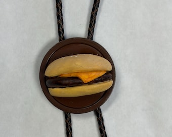 May include: A bolo tie featuring a circular brown base with a miniature hamburger charm attached to the centre. The hamburger has a tan bun, a dark brown patty, and a slice of orange cheese. The cord is black and braided with a textured finish.