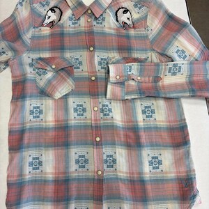 May include: A long-sleeved plaid shirt in shades of pink, blue, and white. The shirt features embroidered opossum heads on the chest and a geometric pattern. It has button closures and two chest pockets. A tag with the letters "EM" is attached to the collar.