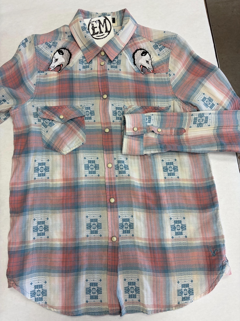 May include: A long-sleeved plaid shirt in shades of pink, blue, and white. The shirt features embroidered opossum heads on the chest and a geometric pattern. It has button closures and two chest pockets. A tag with the letters "EM" is attached to the collar.