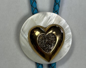 May include: A bolo tie featuring a round, iridescent white mother-of-pearl disc centred with a gold-toned heart ornament. The heart has a textured, dark glittery centre. The cord is made of braided bright turquoise material.