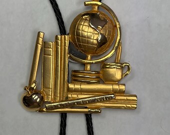 May include: A gold-toned bolo tie featuring a school-themed pendant. The pendant depicts a stack of books, a globe on a stand, a small apple, a ruler, and a coffee mug. The pendant is attached to a black braided cord that loops through the back.