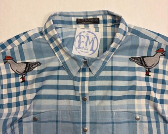 May include: A light blue and white plaid button-down shirt featuring two embroidered pigeons wearing red hats and red boots on the chest. The collar tag reads Ellie Monster Nashville and a white tag with the letters EM is attached to the collar.