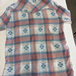 May include: A short-sleeved button-down shirt with a plaid pattern in shades of pink, blue, and white. The shirt features a Western-style yoke and decorative blue geometric designs throughout the fabric.