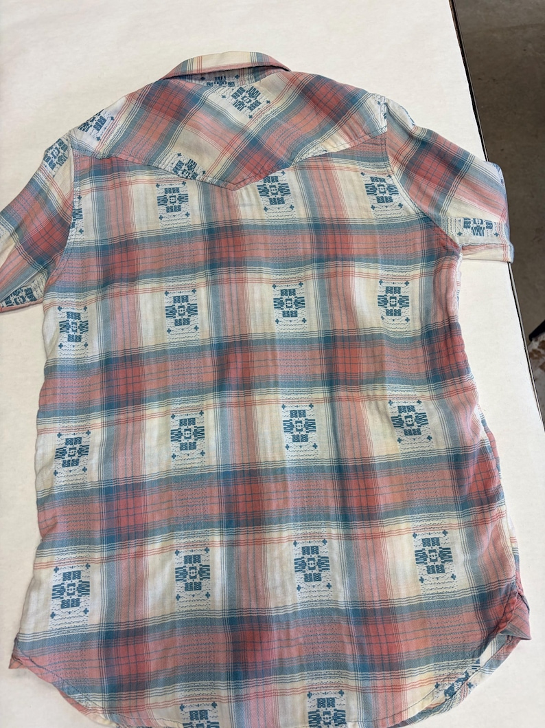 May include: A short-sleeved button-down shirt with a plaid pattern in shades of pink, blue, and white. The shirt features a Western-style yoke and decorative blue geometric designs throughout the fabric.