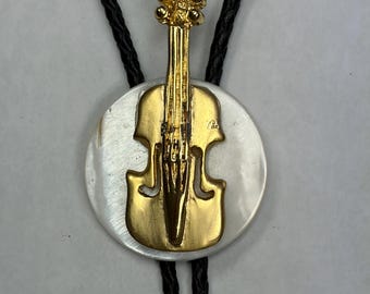 May include: A bolo tie featuring a gold-toned violin pendant mounted on a circular white mother-of-pearl disc. The pendant is attached to a black braided cord that passes through the back of the disc, creating a functional neck accessory.