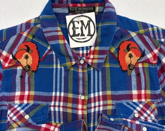 May include: Blue plaid shirt with embroidered bison motifs on the shoulders. The shirt has a classic collar, pearl snap buttons, and the brand name "Ellie Monster Nashville" on the collar.