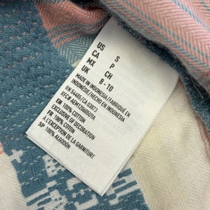 May include: Close-up of a white clothing tag with black text, including size information and fabric details. The tag is on a blue, pink, and white patterned fabric, with the text "100% COTTON".