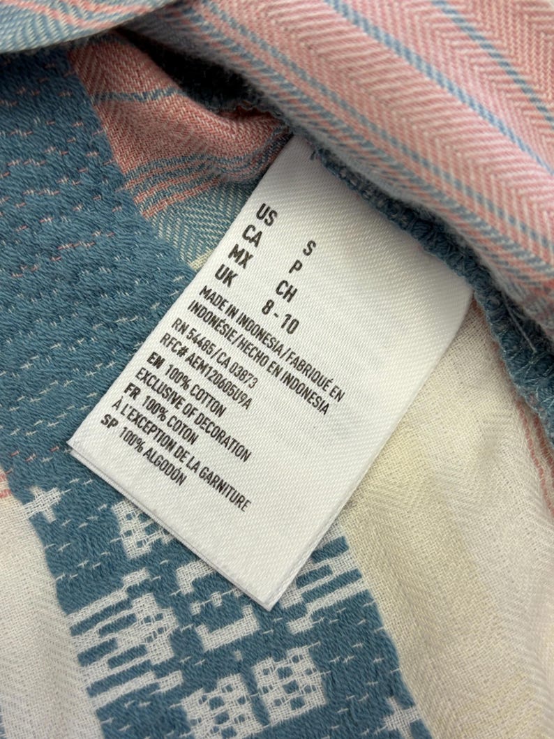 May include: Close-up of a white clothing tag with black text, including size information and fabric details. The tag is on a blue, pink, and white patterned fabric, with the text "100% COTTON".