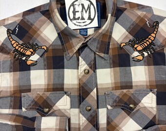 May include: A brown, navy, and cream plaid shirt with button fastenings. The shirt has two embroidered orange scorpions with lightning bolts. The label reads "Ellie Monster Nashville" and "EM".