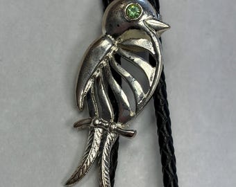 May include: A silver bolo tie with a bird motif and a green gemstone eye. The bird sits on a decorative branch, and the tie has a black braided leather cord.
