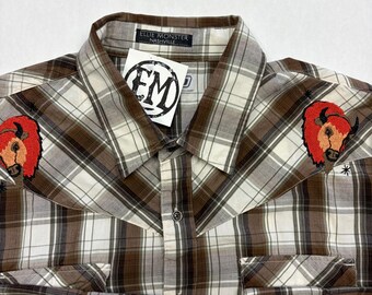 May include: A brown, white, and tan plaid button-up shirt with two front pockets and long sleeves. The shirt features embroidered red bison on the shoulders and a tag that reads "ELLIE MONSTER NASHVILLE".