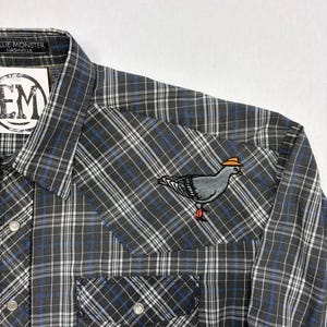 May include: A gray, black, and blue plaid shirt with a pigeon patch on the pocket. The pigeon is wearing a yellow hat and has red feet. The shirt has snap closures and a collar. The label says "BLUE MONSTER NASHVILLE".
