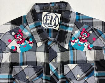 May include: A long-sleeve button-down shirt in a black, white, and blue plaid pattern. The shirt features two chest pockets with flaps and embroidered clown patches on each shoulder. A tag with the text EM is attached to the collar area.